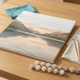 Paint by numbers kit in progress on a wooden table with numbered acrylic paint pots, brushes and a partly painted lake-and-mountain canvas