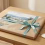 A finished landscape paint by numbers canvas partly wrapped as a gift with a teal ribbon, beside numbered paint pots, a brush and a sprig of eucalyptus on a light wooden table