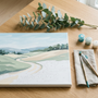 A calm paint by numbers landscape canvas of soft green hills on a wooden table with brushes, teal paint pots and a eucalyptus sprig, illustrating a relaxing painting setup