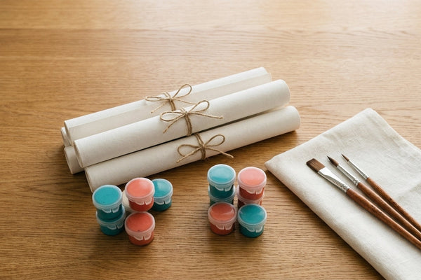 Three rolled blank paint by numbers canvases tied with twine beside teal and coral acrylic paint pots and three wooden brushes on a warm oak table