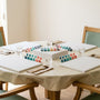 A care home activity table laid out for a group paint by numbers session: four blank canvas boards, sets of teal and coral acrylic paint pots, chunky-handled brushes and water glasses on a wipeable cream cloth in bright daylight