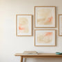 Gallery wall of four light oak frames holding soft abstract watercolour washes above an oak console table with dried grasses and a teal ceramic dish