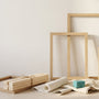 Three empty light oak picture frames leaning against a cream wall with a bundle of wooden stretcher bars and a rolled blank canvas on pale floorboards