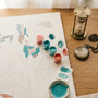 Overhead flat-lay of a partially completed paint by numbers canvas with teal and coral paint pots, a brass hourglass, and a vintage pocket watch on a warm wood desk