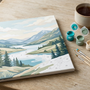 A partly finished paint by numbers landscape canvas on a wooden table with brushes, teal and green paint pots and a cup of tea in calm evening light