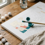 A paint by numbers canvas mid-progress on a wooden desk with a paintbrush resting in a teal paint pot, numbered paint pots and natural window light, evoking a calm, focused painting session