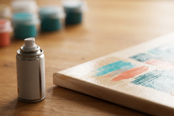 Close-up still life of a varnish spray can beside the glossy, freshly sealed corner of a paint by numbers canvas with abstract teal and coral paint blocks