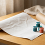 A rolled paint by numbers canvas partially unrolled on a wooden table showing soft travel crease lines, next to teal and coral paint pots and the cardboard shipping tube