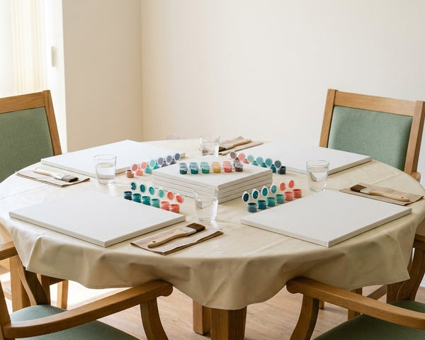 A care home activity table laid out for a group paint by numbers session: four blank canvas boards, sets of teal and coral acrylic paint pots, chunky-handled brushes and water glasses on a wipeable cream cloth in bright daylight