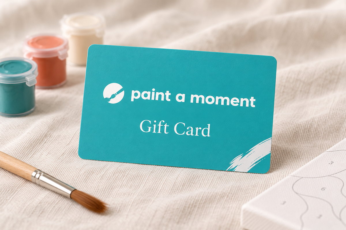 Paint A Moment gift card in teal with the white logo, standing on linen beside paint pots and a brush