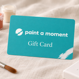 Paint A Moment gift card in teal with the white logo, standing on linen beside paint pots and a brush